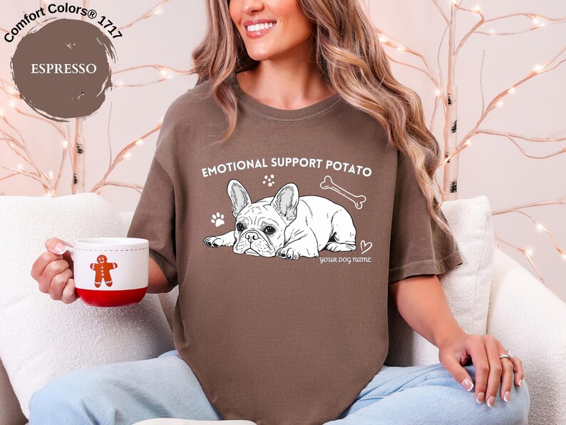 May include: Espresso-coloured t-shirt with the text "EMOTIONAL SUPPORT POTATO" and a graphic of a French Bulldog. The shirt also features a bone, paw prints, and a heart. A white mug with a gingerbread man is held in the hand.