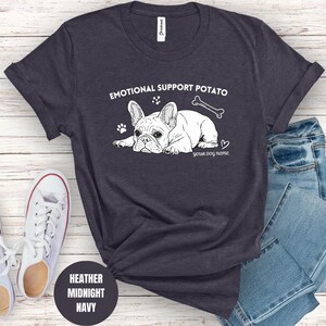 May include: Heather midnight navy t-shirt with a white graphic of a French bulldog, a bone, and the text "EMOTIONAL SUPPORT POTATO". Includes space for a dog's name. White trainers and blue jeans are next to the shirt.
