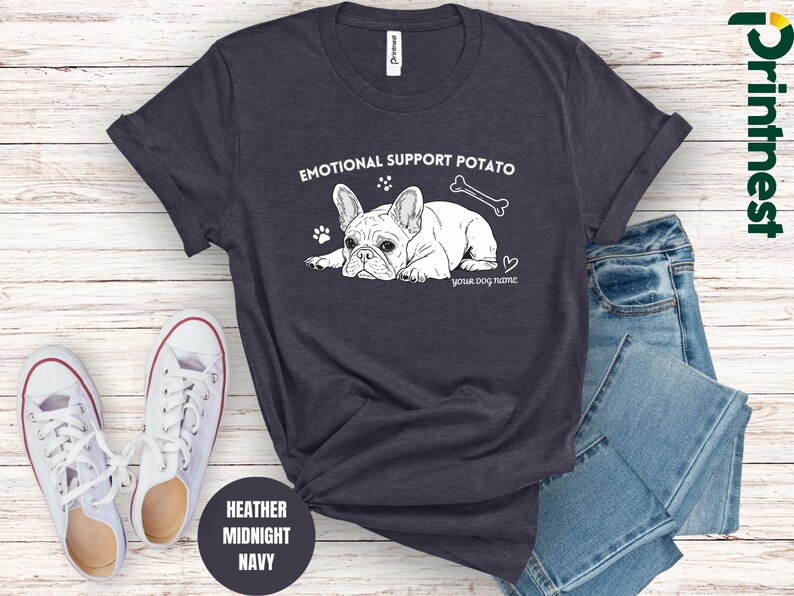 May include: Heather midnight navy t-shirt with a white graphic of a French bulldog, a bone, and the text "EMOTIONAL SUPPORT POTATO". Includes space for a dog's name. White trainers and blue jeans are next to the shirt.