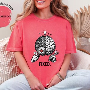May include: A watermelon-colored t-shirt featuring a graphic of a split brain. One side shows a circuit board pattern with a magnifying glass over a red bug, while the other side shows a biological brain. The text 404 and FIXED are printed below.