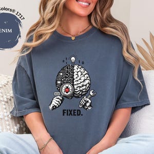 May include: A denim blue short-sleeve t-shirt featuring a graphic of a brain split between a circuit board and organic tissue. One hand holds a magnifying glass over a red bug, while the other holds a wrench. Above is a lightbulb and binary code, with the text FIXED. below.