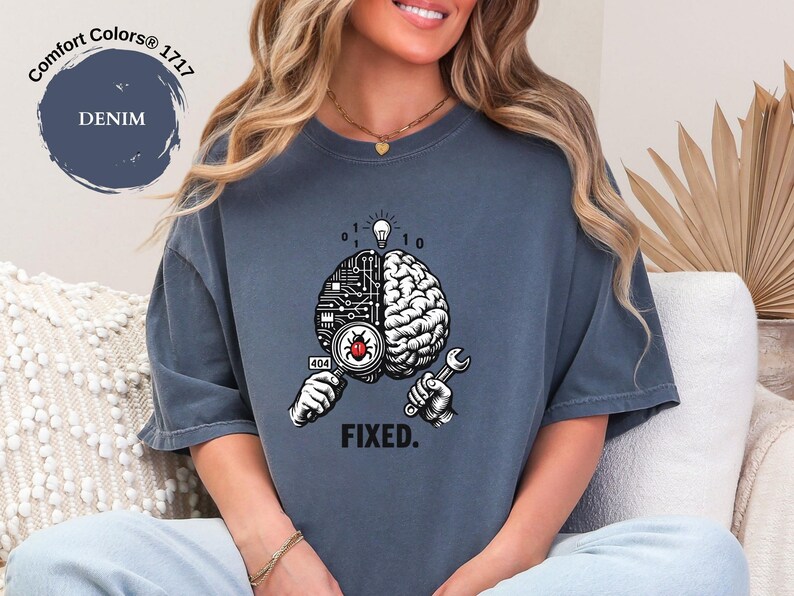 May include: A denim blue short-sleeve t-shirt featuring a graphic of a brain split between a circuit board and organic tissue. One hand holds a magnifying glass over a red bug, while the other holds a wrench. Above is a lightbulb and binary code, with the text FIXED. below.
