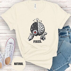 May include: A cream-colored t-shirt featuring a graphic of a brain split between a circuit board and organic tissue. One hand holds a magnifying glass over a red bug with a 404 label, while the other holds a wrench. The word FIXED is printed below.