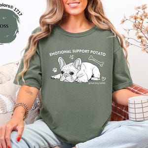 May include: A moss green t-shirt featuring the text "EMOTIONAL SUPPORT POTATO" and a white illustration of a French Bulldog. The design includes a paw print, a bone, and a heart. The words "your dog name" are also included.