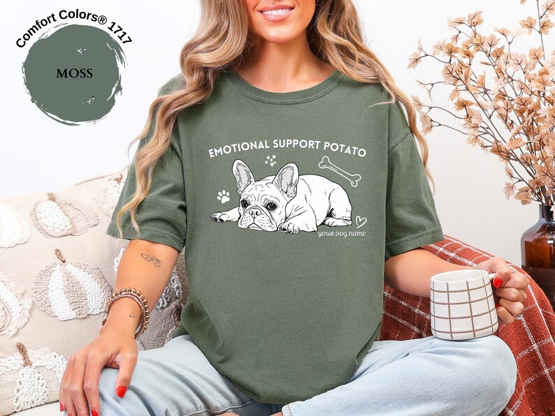May include: A moss green t-shirt featuring the text "EMOTIONAL SUPPORT POTATO" and a white illustration of a French Bulldog. The design includes a paw print, a bone, and a heart. The words "your dog name" are also included.