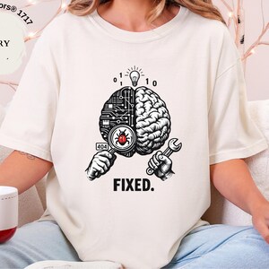 May include: An ivory t-shirt featuring a graphic of a brain split between a circuit board and organic tissue. One hand holds a magnifying glass over a red bug with a 404 label, while the other holds a wrench. A lightbulb and binary code appear above.