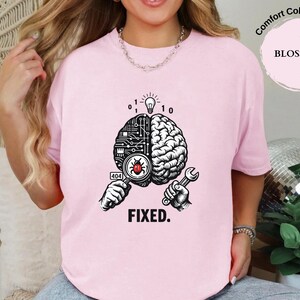 May include: A light pink t-shirt featuring a graphic of a split brain, with one side showing circuit board patterns and the other side showing organic brain tissue. A magnifying glass reveals a red bug, and the text reads 404 and FIXED.