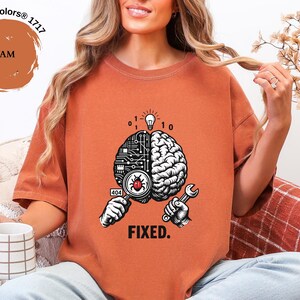 May include: A yam-colored t-shirt featuring a graphic of a brain split between a circuit board and organic tissue. The design includes a magnifying glass over a bug, a wrench, binary code, a lightbulb, and the text "404" and "FIXED."