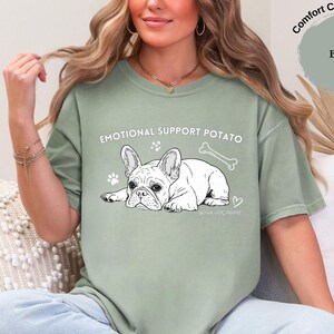 May include: Sage green t-shirt featuring the text "EMOTIONAL SUPPORT POTATO" and a black and white illustration of a French Bulldog. The design includes paw prints, a bone, and a heart.