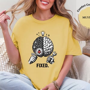 May include: A mustard yellow t-shirt featuring a graphic of a brain split into a digital circuit side and a biological side. A magnifying glass reveals a red bug, a lightbulb glows above, and a wrench is held below with the text FIXED. and 404.