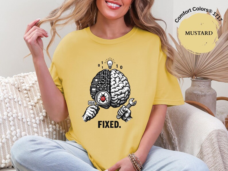 May include: A mustard yellow t-shirt featuring a graphic of a brain split into a digital circuit side and a biological side. A magnifying glass reveals a red bug, a lightbulb glows above, and a wrench is held below with the text FIXED. and 404.