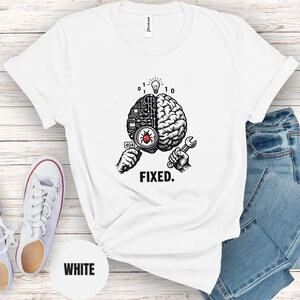 May include: A white t-shirt featuring a black and white graphic of a brain split between a circuit board and organic tissue. One hand holds a magnifying glass over a red bug, while the other holds a wrench. Above is a lightbulb and binary code, with the text FIXED below.