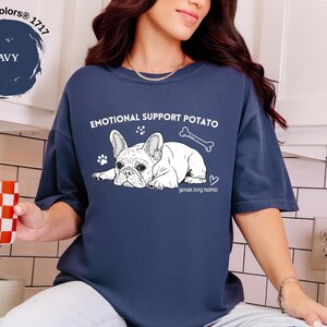 May include: Navy blue t-shirt featuring a white illustration of a French Bulldog, a bone, and the text "EMOTIONAL SUPPORT POTATO". The shirt also includes paw prints and a heart. The text "your dog name" is below the dog graphic.