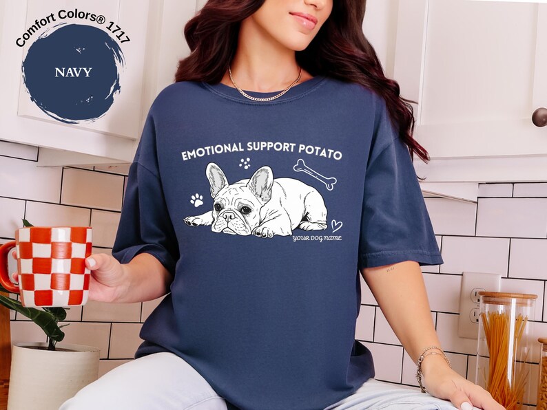 May include: Navy blue t-shirt featuring a white illustration of a French Bulldog, a bone, and the text "EMOTIONAL SUPPORT POTATO". The shirt also includes paw prints and a heart. The text "your dog name" is below the dog graphic.