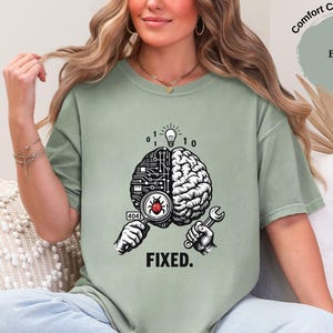 May include: A sage green t-shirt featuring a graphic of a brain split between a circuit board and organic tissue. One hand holds a magnifying glass over a red bug, while the other holds a wrench. Above is a lightbulb and binary code. The text reads FIXED.
