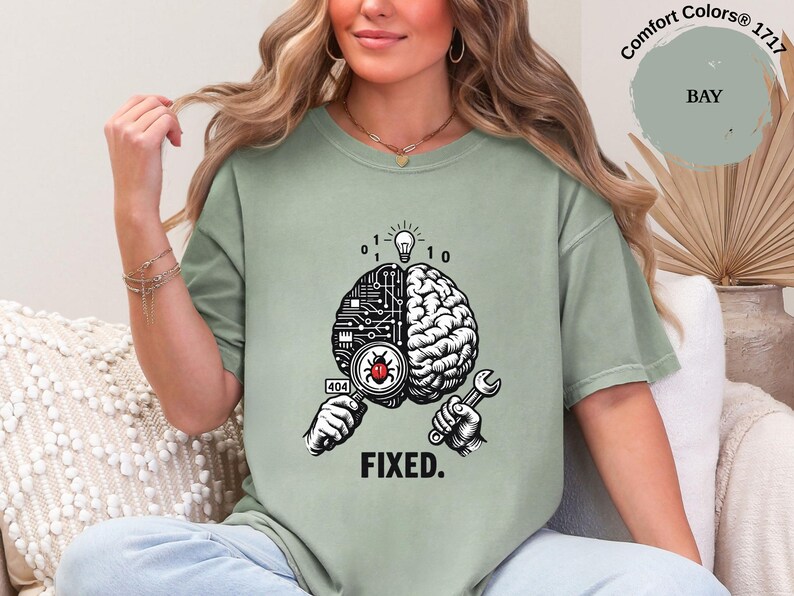 May include: A sage green t-shirt featuring a graphic of a brain split between a circuit board and organic tissue. One hand holds a magnifying glass over a red bug, while the other holds a wrench. Above is a lightbulb and binary code. The text reads FIXED.