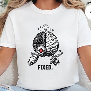 May include: A white t-shirt featuring a graphic of a brain split into a circuit board and organic side. One hand holds a magnifying glass over a red bug with a 404 label, while the other holds a wrench. Above is a lightbulb and binary code, with the text FIXED.