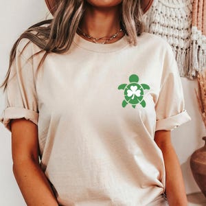 May include: Beige t-shirt featuring a green turtle design with a shamrock on the front. The short-sleeved shirt has rolled-up sleeves and a relaxed fit. The shirt is made of a soft, neutral-colored fabric.