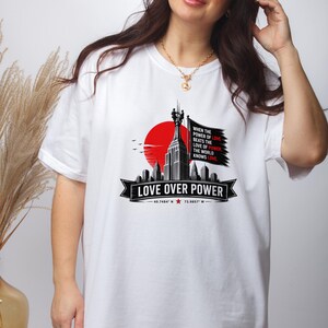 May include: A white t-shirt featuring a graphic of a skyscraper silhouette against a red sun. The design includes a black flag with the text: WHEN THE POWER OF LOVE BEATS THE LOVE OF POWER, THE WORLD KNOWS LOVE. Below is LOVE OVER POWER.