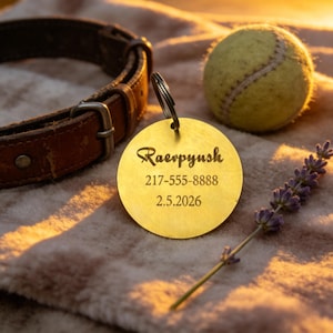 May include: A circular gold pet tag engraved with the name "Raevpyush", a phone number, and a date. A brown leather dog collar, a tennis ball, and lavender are also in the image.