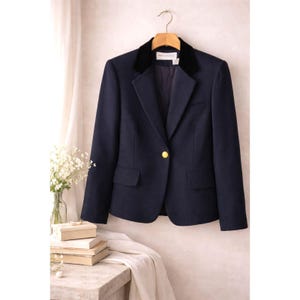 Puede incluir: Una blazer azul marino con cuello de terciopelo negro y un solo botón dorado. La blazer está colgada de una percha de madera. La blazer tiene dos bolsillos delanteros.