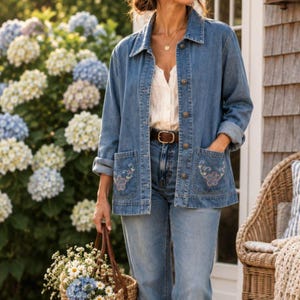 May include: A blue denim jacket featuring a classic collar, long sleeves, and two front pockets with delicate floral embroidery. The jacket is worn over a white button-down shirt and blue denim jeans, secured with a brown leather belt.