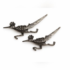 May include: Two metal roadrunner bird pins. The birds are in a running pose, with detailed wings and tail feathers. The pins are a dark silver color and have a textured appearance, with a small cluster of embellishments on the head and back.