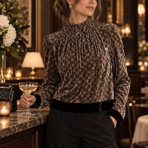 May include: A long-sleeved blouse featuring a high, gathered neckline and a repeating black monogram pattern on a metallic bronze fabric. The garment includes a solid black velvet hemline and matching black velvet cuffs on the sleeves.