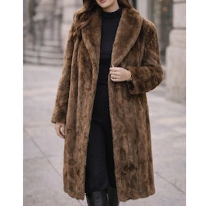 May include: A long, brown faux fur coat with a wide collar. The coat is knee-length and has a textured appearance. The person is wearing a black turtleneck and black pants. The coat is a stylish outerwear option.