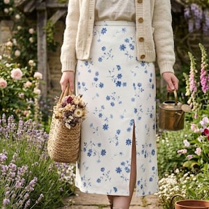 May include: A woman wearing a cream-colored cardigan and a white skirt with a blue floral pattern. She is holding a woven basket filled with dried flowers and a small brass watering can. The skirt has a front slit.