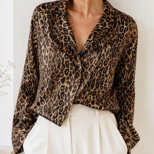 May include: A long-sleeved button-down shirt with a leopard print pattern in shades of brown and black. The shirt is paired with white pleated trousers. The top has a classic collar and a relaxed fit, suitable for various occasions.
