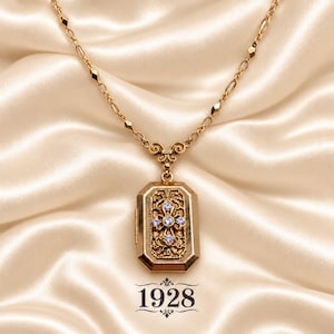 May include: A gold-tone locket necklace featuring a rectangular pendant with an ornate filigree center and four small clear crystals arranged in a cross pattern. The pendant hangs from a delicate gold-tone chain with small geometric accents.
