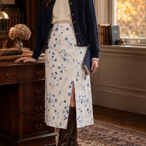 May include: A high-waisted midi skirt in white fabric featuring a scattered blue floral pattern and a vertical front slit. The skirt is paired with a cream-colored button-down shirt, a dark navy blue cable-knit cardigan, and dark brown boots.