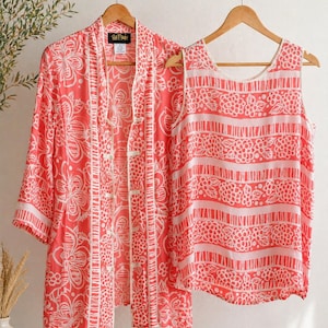 May include: Coral-colored, floral-patterned top and matching open-front jacket hanging on a wooden rack. The sleeveless top features horizontal stripes with floral accents. The jacket has a button-up front and three-quarter sleeves. The set is made of a lightweight fabric.