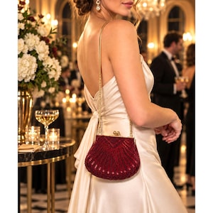 Puede incluir: Un bolso de mano de noche en forma de concha de color rojo intenso, con intrincados abalorios y un marco de metal dorado con cierre de broche. El bolso incluye una fina correa de cadena de metal dorado y tiene un diseño estructurado en abanico.