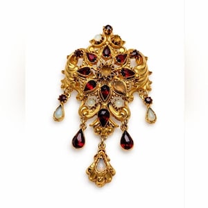 May include: Ornate gold-tone brooch with a floral design, featuring red and white gemstones. The brooch has a central starburst design and teardrop-shaped dangles. A vintage-inspired jewelry piece.