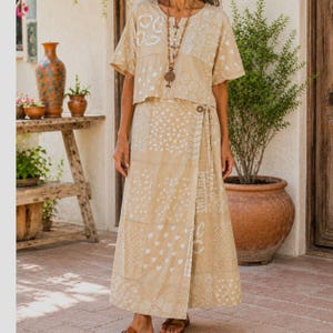 May include: A beige two-piece set consisting of a short-sleeved top and a wrap-style skirt, both featuring a white block-printed pattern of circles, hearts, and geometric shapes. The top has a round neckline and a long pendant necklace is worn.