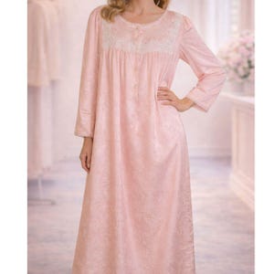 May include: A long, light pink nightgown with a floral pattern. The nightgown features long sleeves, a round neckline with white lace trim, and a button-down front. The fabric appears soft and flowing, suitable for sleepwear.