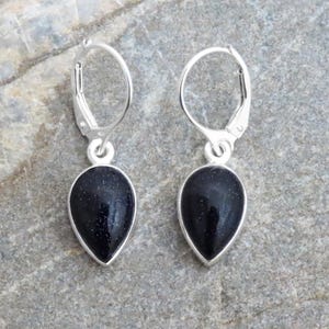 Könnte beinhalten: Ein Paar silberfarbene Ohrringe mit einem tropfenförmigen, dunkelblauen Stein. Die Ohrringe haben einen Klappverschluss und werden auf einer grauen Steinoberfläche präsentiert. Schmuck.