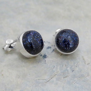Op de afbeelding: Een paar oorstekers met een zilveren zetting. Elke oorbel heeft een ronde, donkerblauwe steen met sprankelende insluitsels. De oorbellen worden gepresenteerd op een lichtgrijze ondergrond.