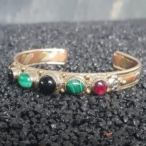 Könnte beinhalten: Ein silbernes Armreifarmband mit eingelegten Edelsteinen. Das Armband hat grüne Malachit-, schwarzen Onyx- und roten Granatsteine. Die Metallarbeiten haben ein Mischmetalldesign mit Gold- und Kupferakzenten.