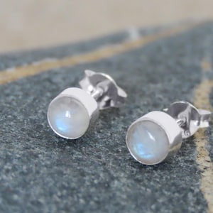 Könnte beinhalten: Ein Paar silberne Ohrstecker mit runden, irisierenden Mondstein-Cabochons. Die Ohrringe sind in einer einfachen Lünettenfassung gefasst, und die Stifte haben einen Schmetterlingsverschluss. Die Ohrringe werden auf einer strukturierten grauen Oberfläche präsentiert.