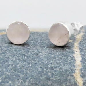 Könnte beinhalten: Ein Paar Ohrstecker mit runden, hellrosa Edelsteinen, die in Silber gefasst sind. Die Ohrringe werden auf einer strukturierten, grauen Oberfläche gezeigt, die ihr schlichtes, elegantes Design hervorhebt. Diese Ohrringe sind ein klassisches Schmuckaccessoire.