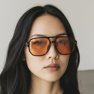 May include: Black square-framed sunglasses with orange-tinted lenses. The sunglasses have a double bridge and a thick frame. The person in the image is wearing a black top.