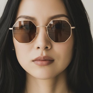 May include: Close-up of a person wearing gold-rimmed, octagonal sunglasses with brown lenses. The glasses have a geometric shape and a thin metal frame. The person has dark hair and is looking forward.