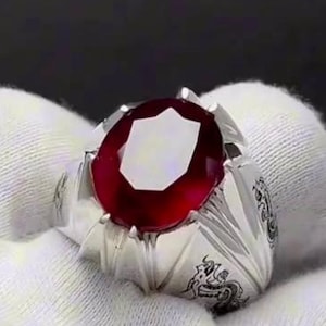 May include: A silver ring with an oval, deep red gemstone. The ring has intricate carvings on the band and is set against a white background. The gemstone is the focal point, catching the light.