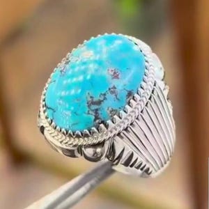 May include: A silver ring with a large, oval turquoise stone. The stone is a vibrant blue with brown veining. The ring has a detailed, textured band and a decorative setting around the stone. The ring is held by a pair of tweezers.