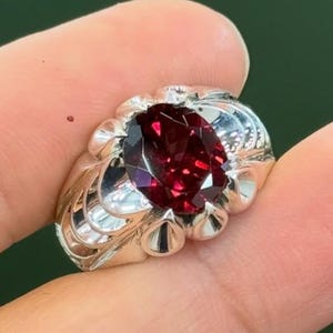 May include: A silver ring with a large, oval-cut, deep red gemstone as the centerpiece. The ring has a detailed, ornate design around the gemstone and on the band. The ring is displayed against a green background. The logo "MK Dimonds Jewelery" is in the bottom right corner.