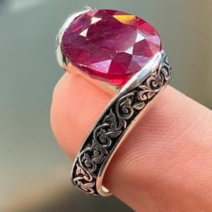 May include: A silver ring with an oval, faceted ruby gemstone. The ring band has an ornate, engraved design. The ruby is a deep red colour, and the ring is held up by a finger. The logo "MK Dimonds Jewelery" is in the bottom right corner.