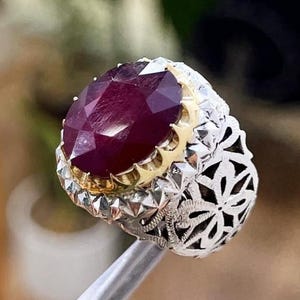 May include: A silver ring featuring a large, oval-cut ruby gemstone. The ruby is set in gold and surrounded by a detailed silver filigree band. The ring is held by tweezers. The ring is a piece of jewellery.